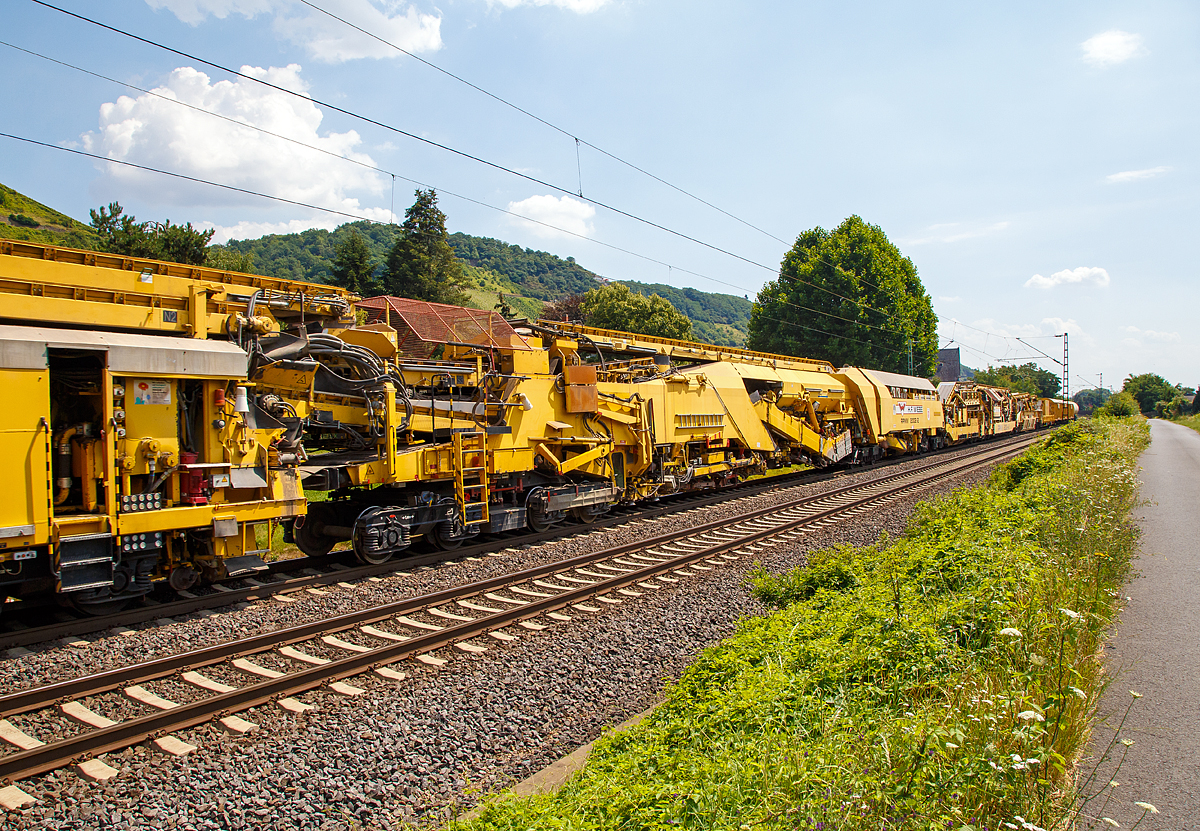 
Die Wiebe Recycling-Planumsverbesserungsmaschine mit Schotterrecycling RPMW 2002-2 am 14.07.2018 im Zugverband auf der Fahrt durch Leutesdorf,  auf der KBS 465 (Rechte Rheinstrecke) in Richtung Norden. 

Über 140 Meter lang, knapp 5 Meter hoch und gute 560 Tonnen schwer – die Gleisbaumaschine RPMW 2002-2 gehört zu den größten der Welt. Mit ihrer beachtlichen Leistung und dem umfänglichen Leistungsspektrum ist sie ein äußerst wichtiger Bestandteil des Maschinenparks der Unternehmensgruppe Wiebe.

Die Recycling Planumsverbesserungsmaschine führt verschiedene Gleisbauarbeiten durch – und das mit einer Motorengesamtleistung von über 2100 Kilowatt. Diese Kraft kommt bei Totalsanierungen zum Tragen, bei denen die RPMW 2002-2 mehrere Arbeitsschritte simultan ausführt: Die verschmutzte Schotterbettung wird ausgebaut, das abgetragene Bettungsmaterial aufbereitet, Schotter recycelt und die Unterbauplanumsschicht erneuert.

TECHNISCHE DATEN:
Hersteller: Plasser & Theurer
Baujahr, Erstzulassung: 2000
Spurweite: 1.435 mm
Anzahl der Achsen: 29, davon 12 angetrieben
Baumaschinennummer: Aushubmaschine 3369, Recyclingmaschine 3368, Antriebswagen 3367,  Stopf- u. Einschotterungsmaschine 3370
Motoren: Caterpillar 
Leistung: 2.860 PS
Gesamtlänge über Puffer: 140,90 m
Breite: 3,29 m
Höhe: 4,65 m
Gewicht: 560 t
V zul. im Zug eingestellt: 100 km/h
V max. bei Eigenfahrt: 20 km/h
Fahrradius: 120 m

Der große Unterschied zwischen Planumsverbesserungsmaschine und Bettungsreinigungsmaschinen besteht darin, dass die Planumsverbesserungsmaschine nicht nur den Schotter ausheben und recyceln kann, sondern auch einen Teil des Planums (Unterbau) ausheben (bis 1.200 mm unter SOK). Zudem kann die RPM vor der Neuschotterung (Recycling- mit Neumaterial) auch eine Planumsschutzschicht (PSS-Material) einbringen.
Der Neuschottern wird durch die Stopfeinrichtung 09-32 Stopfsatellit im Gleisbett verdichtet und das Gleis ist danach wieder mit 70 km/h befahrbar.

Hier in der Bildmitte die Planumsaushubkette:
Hat die Schotteraushubkette die Schotterschicht abgetragen, nimmt die Planumsaushubkette das verbleibende Boden- und Bettungsmaterial bis zum Erdplanum auf. Auch diese Aushubkette lässt sich stufenlos justieren, nämlich auf die gewünschte Räumbreite, Räumtiefe und Planumsquerneigung. Erneut transportieren die Förderanlagen den Aushub zur Maschinenfront, wo er auf Materialförder- und Siloeinheiten verladen wird.