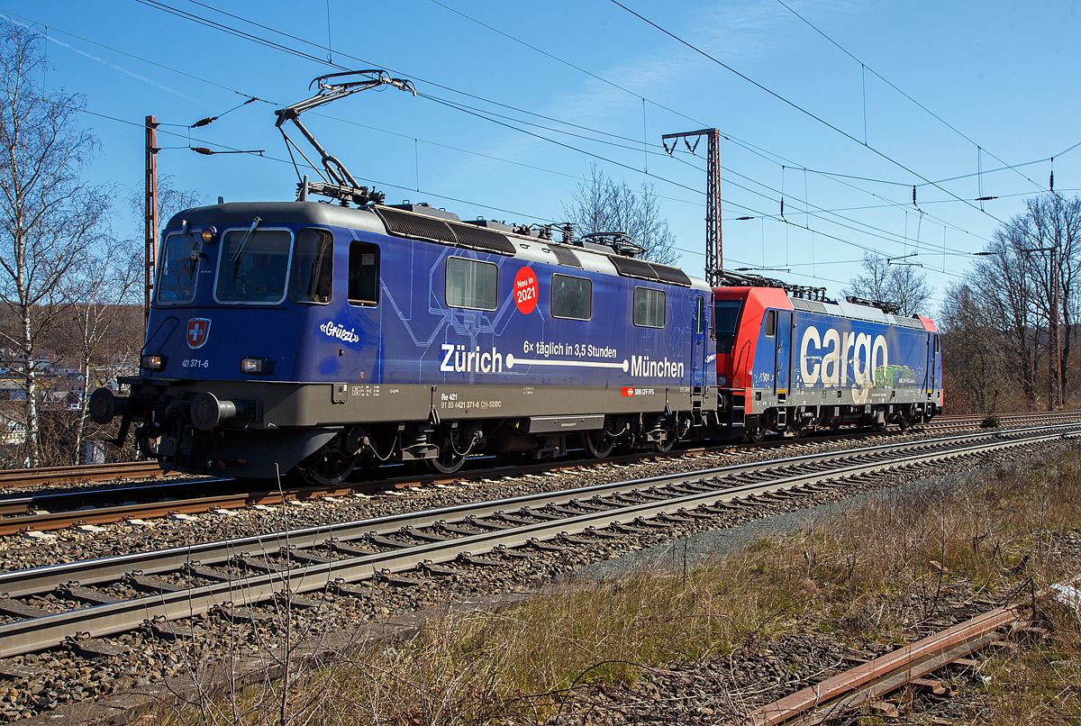 Ja, „Gr�ezi“ oder „Servus“.....
Die SBB Cargo Re 421 371-6 (91 85 4421 371-6 CH-SBBC), ex Re 4/4 II 11371, mit Werbung Z�rich – M�nchen 6 x t�glich in 3,5 Stunden, f�hrt am 30.03.2021 mit der SBB Cargo Re 484 016-1(91 85 4484 016-1 CH-SBBC), ex E 484.016 SR, einer TRAXX F140 MS am Haken durch Rudersdorf (Kr. Siegen), an der Dillstrecke (KBS 445), in Richtung Siegen.

Eine SBB Cargo Re 484 wie die 016-1 kann man eigentlich Deutschland nur mit viel Gl�ck sehen, denn sie haben nur die Zulassungen f�r die Schweiz und Italien. Sie haben keinen Stromabnehmer mit breiterer Wippe gem�� deutscher Vorschrift, auch die die deutsche Zugsicherung Indusi fehlt ihnen. Der Lz war auf dem weg nach Krefeld.

Lebensl�ufe:
Die Re 421 371-6 wurde 1983 von der SLM Winterthur (Elektrik BBC/ SAAS) unter der Fabriknummer 5235 gebaut und als Re 4/4 II 11371 an die SBB geliefert. Im Jahr 2002 erfolgte der Umbau zur Re 421 f�r die SBB Cargo (Einsatz auch in Deutschland). Die zugelassene H�chstgeschwindigkeit f�r die Schweiz betr�gt 140 km/h und f�r Deutschland 120 km/h.

Die Re 484 016-1 eine Bombardier TRAXX F140 MS wurde 2004 von Bombardier in Kassel unter der Fabriknummer 34009 gebaut und an die SBB Cargo AG, als E 484.016 SR geliefert. 