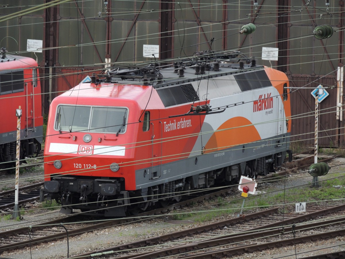 Mit Tele so gut es ging durch viele Fahrdrähte durch: 120 112-8 mit Märklin-Werbung vor dem Lokschuppen in Ulm am 19.05.2012.