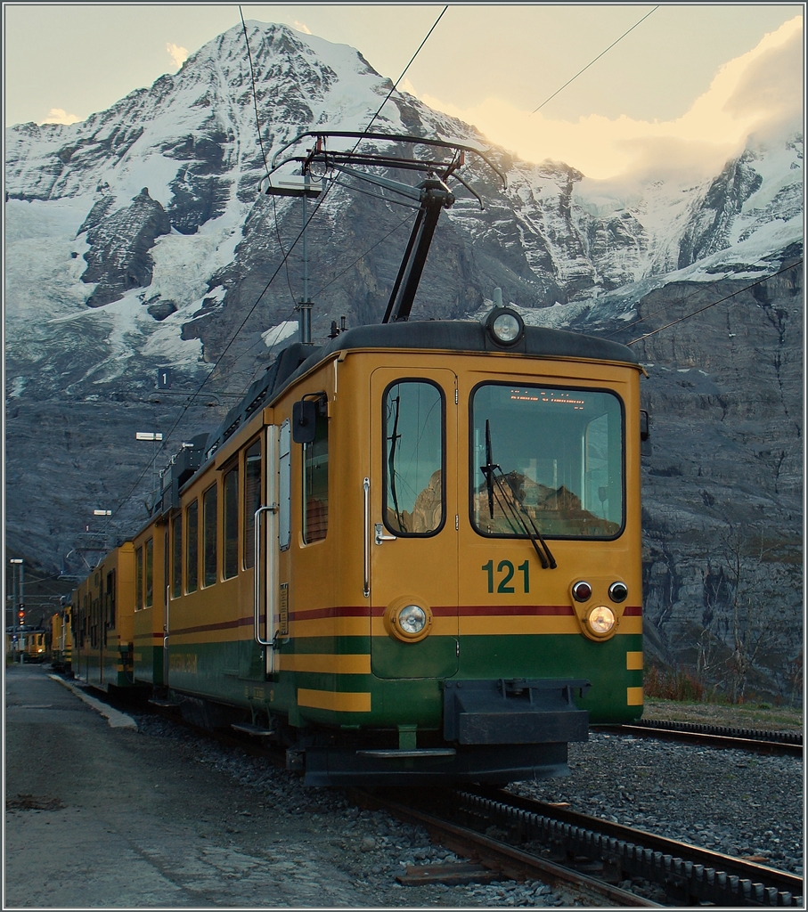 Weiterhin mit einem Stück  fehlender  Fahrleitung, nun aber farblich verändert, präsentiert sich der WAB BDeh 4/4 121 vor dem Mönch beim Halt auf der noch schattigen Wengern Alp. 
9. Okt. 2014