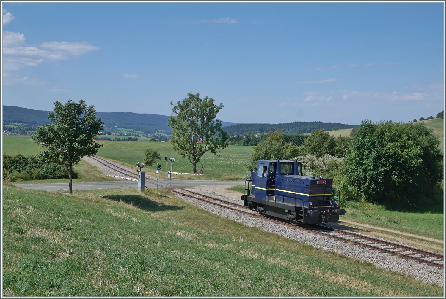 30 ANS CONI'FER /30 Jahre Coni'Fer - Nachdem der Coni'Fer locotracteur Tm 2/2 (ex TSOL Tm 2/2 238 291-9) die Coni'Fer  Orient Express  in Fontaine Ronde rangiert hat, wird die Lok wohl für das Manöver in Les Hôpitaux Neufs gebraucht, jedenfalls ist sie bei Le Touillon auf dem Weg dorthin. Das Bild zeigt auch einen Blick auf den Bahnübergang mit der Strasse D 45 und den entsprechenden Signalen und Ampeln für Strasse und Schiene.

15. Juli 2023