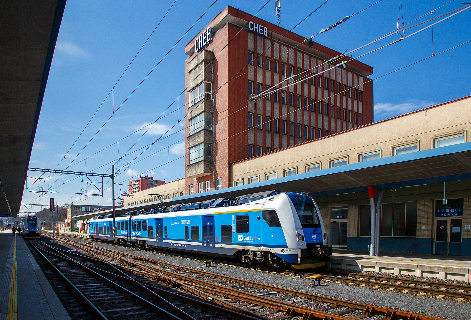 Der Bahnhof Cheb (Eger) am 21.04.2023, hinten (auf Gleis 1) steht der RegioPanter 651 204-0 / 650 204- 1 (CZ-ČD 94 54 1 651 204-0 / CZ-ČD 94 54 1 650 204-1), ein zweiteiliger Zweisystem-Elektrotriebzug (3 kV DC und 25 kV, 50 Hz~) vom Typ �koda 15 Ev, der Česk� dr�hy (ČD) / Plzeňsk� Kraj (deutsch Pilsner Region) als Sp (Spě�n� vlak / Eilzug) nach Pilsen (Plzeň) zur Weiterfahrt bereit. Er hatte uns von Karlovy Vary (Karlsbad) nach Cheb gebracht. 

Links hinten am Gleis 2 steht der RegioPanter 651 203-2 / 650 203-3 (CZ-ČD 94 54 1 651 203-2  / CZ-ČD 94 54 1 650 203-3) als Gegenzug von Pilsen nach Karlsbad.
