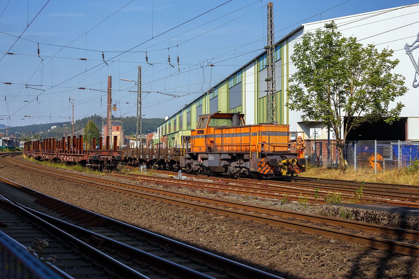 Die DEW 7 (Lok 7 der Deutschen Edelstahlwerke GmbH), zuvor Edelstahlwerke Südwestfalen, rangiert am 05 September 2023 mit vierachsigen Drehgestellflachwagen am Haken, vom Anschlussgleis zurück aufs Werksgelände in Siegen-Geisweid.

Die Lok ist eine MaK G 1203 BB und wurde1986 unter der Fabriknummer 1000808 in Kiel gebaut und an die Krupp Stahl AG, Werk Siegen-Geisweid, geliefert.

Die Lokomotive MaK G 1203 BB ist eine dieselhydraulische Lokomotive, die von der Maschinenbau Kiel (MaK) gebaut wurde, die Achsfolge ist B’B’. Sie erreicht eine maximale Geschwindigkeit von bis zu 70 km/h. Eingebaut wurden Motoren von MTU (745 kW) und für sechs Exporte nach Gabun mit amerikanischen Cummins-Motoren (761 kW). Die MaK G 1203 BB wurde zwischen 1982 und 1991 in 25 Exemplaren gebaut. Sie gingen an 14 verschiedene Eisenbahnbetreiber in Deutschland. Eine Lok ging nach Schweden und sechs nach Gabun.

Im Deutschen Fahrzeugeinstellungsregister wurde für diese Bauart die Baureihennummer 98 80 0273 vergeben.

Ab 1978 wurden im dritten Typenprogramm zwei verschiedene vierachsige Loks angeboten. Die leistungsschwächere erhielt die Bezeichnung G 1201 BB. Sie setzte sich nicht so recht durch und wurde nur einmal gebaut. Als ab 1981 die leistungsstärkeren Motoren der Reihe 396 zur Verfügung standen, änderte man die Typenbezeichnung in G 1203 BB. Äußerlich kann man diese beiden Typen an ihrem Lüftungsgitter an der Vorderseite der langen Haube erkennen.

TECHNISCHE DATEN:
Spurweite: 1.435 mm (Normalspur)
Achsfolge: B´B´
Länge über Puffer 12.500 mm
Drehzapfenabstand: 5.800 mm
Achsabstand im Drehgestell: 2.400 mm
Treibraddurchmesser: 1.000 mm (neu)
größte Breite: 3.100 mm
größte Höhe über Schienenoberkante: 4.220 mm
kleinster befahrbarer Gleisbogen: 60 m
Dienstgewicht: 88 t
Kraftstoffvorrat: 2.500 l
Motor : MTU, Typ 8V 396 TC 13, ein V8-Zylinder-Dieselmotor mit Abgasturbolader und Ladeluftkühlung,
Leistung: 745 kW (1.012 PS)
Drehzahl: 1.800 U/min
Getriebe: Voith, Typ L4r4zU2

Die Deutschen Edelstahlwerke sind die deutsche Produktionsgesellschaft der Swiss Steel Group mit Hauptsitz in Luzern, Schweiz, einem der weltweit führenden Produzenten von Spezialstahl-Langprodukten. Dank des ausschließlichen Einsatzes von Stahlschrott in Elektrolichtbogenöfen zählt die Gruppe zu den europaweit relevantesten Unternehmen in der Kreislaufwirtschaft und ist im Bereich nachhaltig produziertem Stahl – Green Steel – unter den Marktführern. Swiss Steel Group verfügt über eigene Produktions- und Distributionsgesellschaften in über 30 Ländern. 

Die Eckdaten der Geisweider Eisenwerke (heute Teil der Deutschen Edelstahlwerke) :
1845: Gründung des Stahlwerks durch J.H. Dresler als Eisen- und Walzwerk
1872: Errichtung von Hochofen 1
1879: Umbennung in  Geisweider Eisenwerke AG 
1889: Inbetriebnahme des ersten Siemens-Martin-Stahlwerks
1902: Errichtung von Hochofen 2 (mit der noch existierenden Gießhalle)
Ende 1. Weltkrieg: Stillegung beider Öfen (mehrjährige Pause wegen Weltwirtschaftskriese und Quotenvorgaben für Stahlunternehmen)
1920: Aufkauf durch Thyssen und Klöckner, anschließend Teil der Vereinigten Stahlwerke AG, dem zweitgrößten Stahlproduzenten weltweit
1938: Hochofen 2 wird renoviert und modernisiert und geht wieder in Betrieb, Stillstand am Ende des 2. Weltkriegs
1948: der verbliebene Hochofen geht wieder in Betrieb
1951: auf Annordnung der Alliierten Hohen Kommission erfolgte die Zerschlagung der VESTAG und Geisweid Teil der Stahlwerke Südwestfalen AG im Verbund mit dem Stahlwerk Hagen (vorm. Eicken), den Stahlwerken Brünninghaus und der FriedrichThomée AG aus Werdohl
1972: 100 Jahre Hochofenbetrieb in Geisweid, 70 Jahre Hochofen 2 
1974: Übernahme durch Krupp (Krupp Edelstahlprofile)
1978: Installierung eines 140 t Elektrolichtbogenofen (der größte in Deutschland)
1979/1981: Stillegung/Abbruch des Hochofens 2, es war der letzte Siegerländer Hochofen
2004: das Geisweider Werk geht an die Deutschen Edelstahlwerke in der Schmolz&Bickenbach-Gruppe zur Swiss Steel Group gehörend, wieder unter dem Namen Deutsche Edelstahlwerke Specialty Steel. 

Das Stahlwerk in Siegen-Geisweid betreibt einen 140 t Elektrolichtbogenofen und eine 6-adrige Stranggießanlage, um Edelstahl-Knüppel für das eigene Stabstahlwalzwerk und die Drahtstraße in Hagen herzustellen. Der Edelstahl wird aus Schrott hergestellt. Von einstmals rund  7.000 Arbeitsplätzen sind heute noch rund 1.100 verblieben.
