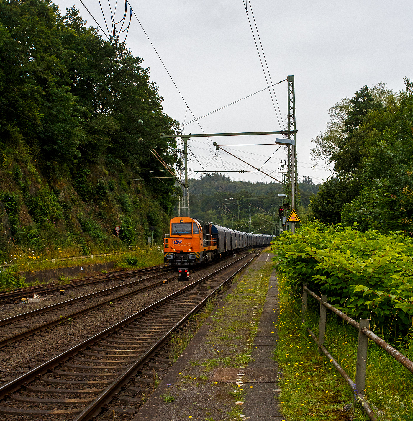 Die KSW 43 (92 80 1273 018-2 D-KSW), die asymmetrische Vossloh G 2000 BB der KSW (Kreisbahn Siegen-Wittgenstein) fährt am 03 Juli 2024, mit einem beladen Coilzug (16 Stück VTG Coilwagen der Gattung Shimmns-ttu, VTG Bez. F46.520D) vom Hafen Duisburg kommend, durch den Bahnhof Scheuerfeld(Sieg) in Richtung Herdorf via Betzdorf. 

In Betzdorf (Sieg) geht es dann über KBS 462 „Hellertalbahn“ nach Herdorf zum Rbf der KSW (Betriebsstätte FGE -Freien Grunder Eisenbahn). Für die Weiterfahrt auf den Pfannenberg (in Neunkirchen-Salchendorf) über nimmt dort dann die KSW 47 (92 80 1271 027-5 D-KSW), ex D 2 der HFM, eine Vossloh G 1000 BB, den Zug.
