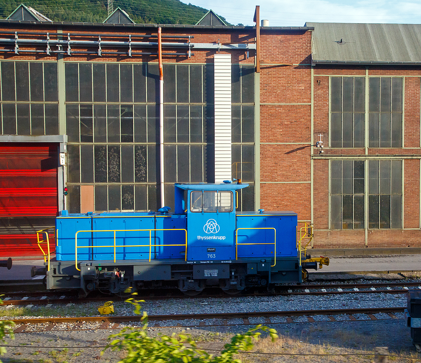 Die Lok 763 der thyssenkrupp Hohenlimburg GmbH, ex 763 Hoesch Hohenlimburg GmbH, ex SK 32 (Siegener Kreisbahn GmbH), am 31.05.2022 auf dem Werksgelände in Hagen-Hohenlimburg, aus einem fahrenden Zug heraus fotografiert.

Die Lok ist eine MaK G 763 C, sie wurde 1990 von der Maschinenbau Kiel GmbH (MaK) unter der Fabriknummer 700099 gebaut und an die damalige Siegener Kreisbahn GmbH als SK 32 geliefert. Zum 23.12.2004 nach der Umbenennung zur Kreisbahn Siegen-Wittgenstein GmbH wurde sie zur KSW 32 und war von 2005 bis 2007 an die MEG - Märkische Eisenbahn-Gesellschaft mbH in Lüdenscheid vermietet. Im Dezember 2007 ging sie über Vossloh Locomotives an die Hoesch Hohenlimburg GmbH als Lok 763, zum 01.07.2016 wurde Hoesch von thyssenkrupp übernommen.