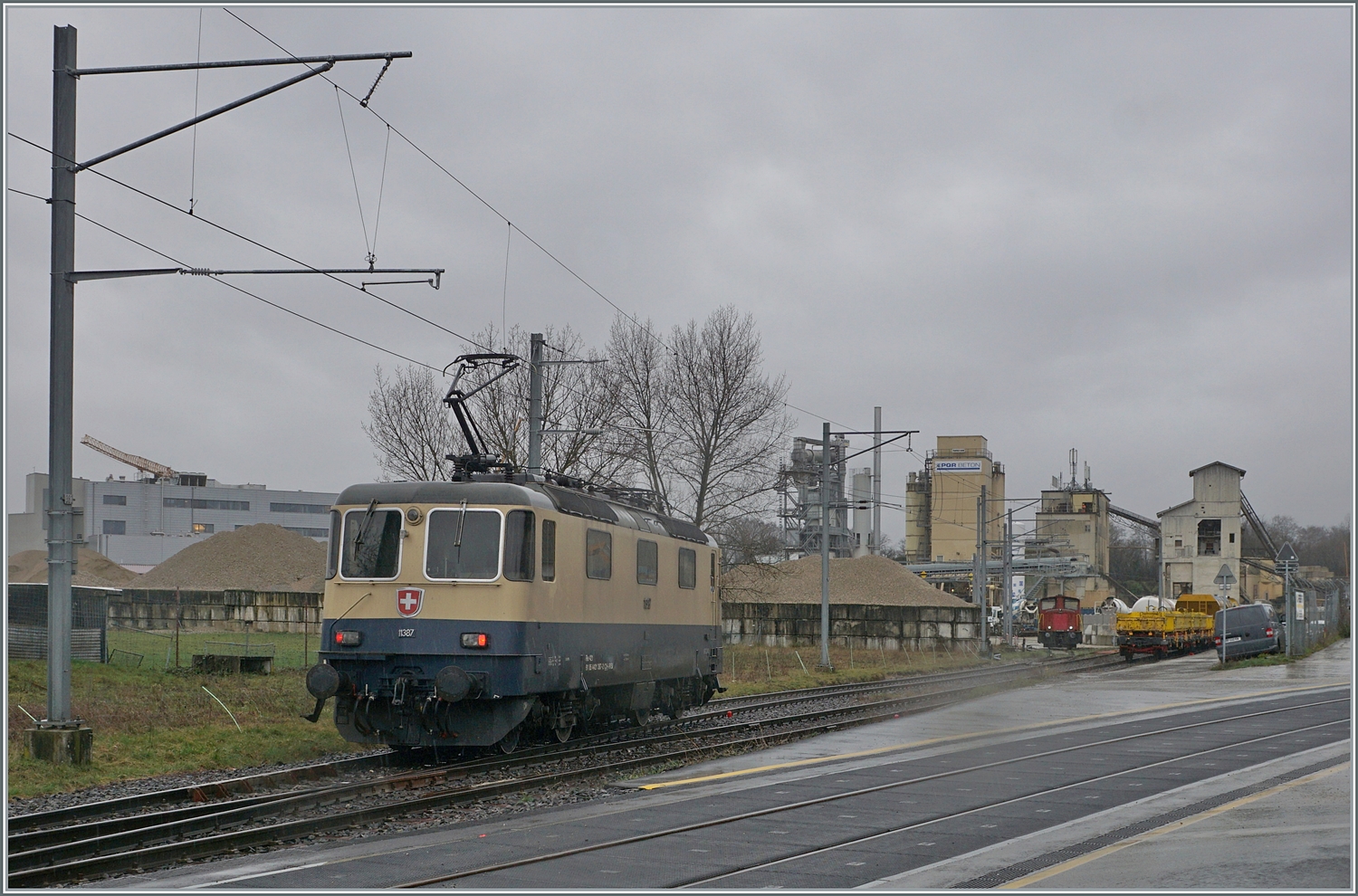Die von Morges gekommene IRSI/IGE  Rheingold  Re 4/4 II 11387 (Re 421 387-2) hat in Gland die Strecke nach Genève verlassen und fährt nun über ein Industriegleis in Richtung Kieswerk, um dort ihren Zug nach Apples abzuholen. 
Die angemietete IRSI/IGE Re 4/4 II 11387 (Re 421 387-2) ersetzt die zur Zeit in der Revision in Bellinzona weilende BAM MBC MBC Re 420 506. 

22. Feb. 2024