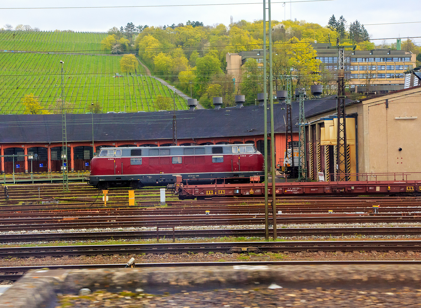 Die V 270.10 „Detlef T.“ alias 221 124-1 (92 80 1 221 124-1 D-SGL) der SGL - Schienen Güter Logistik GmbH (Dachau) steht am 17.04.2023 in Würzburg vor dem Ringlokschuppen, aufgenommen aus einem Zug heraus. 

Die V 270.1 wurde 1964 von der Krauss-Maffei AG in München-Allach unter der Fabriknummer 19244 gebaut und als DB V 200 124 an die Deutsche Bundesbahn geliefert.

Bezeichnungen und Eigentümer waren:
• 1964-1968: DB V 200 124 (Deutsche Bundesbahn)
• 1968-1989: DB 221 124-1 (Deutsche Bundesbahn)
• 1989-2002: ΟΣΕ A 419 (OSE- Organismós Sidirodrómon Elládos / griechische Eisenbahn)
• 2002-2011: 221 124-1 (PE - Prignitzer Eisenbahn GmbH)
• seit 2011: V 270.10 „Detlef T.“ alias 221 124-1 (92 80 1 221 124-1 D-SGL) der SGL - Schienen Güter Logistik GmbH, Dachau

Bei der DB-Baureihe V 200.1 handelt es sich um eine Diesellokomotive der Deutschen Bundesbahn. Von 1962 bis 1965 wurden insgesamt 50 Lokomotiven beschafft, ab 1968 wird sie als Baureihe 221 geführt.  Die Baureihe V 200.1 ist eine Weiterentwicklung der Baureihe V 200.0. Von der Baureihe V 200.0 unterscheidet sich die V 200.1 im Wesentlichen durch die stärkeren Antriebsmotoren von 2 × 1.350 PS gegenüber 2 × 1.100 PS. Die Höchstgeschwindigkeit beträgt 140 km/h. Diese wurden notwendig, da durch das gestiegene Verkehrsaufkommen die Züge länger und schwerer geworden waren. Dadurch kamen die Lokomotiven der Baureihe V 200.0 an ihre Leistungsgrenzen, was nicht selten zu Verspätungen führte. Das höhere Gewicht der neuen Motoren wurde zum Teil durch den Einsatz von Leichtbaustoffen und die Verwendung eines leichteren Kessels für die Dampfheizung wieder ausgeglichen. Die Leichtbauweise führte zu Beginn der 1980er Jahre zu zunehmenden Korrosionsschäden. Der hohe Unterhaltungsaufwand der zweimotorigen Lokomotiven und der rückläufige Güterverkehr im Ruhrgebiet führte bis Ende Mai 1988 zur Ausmusterung der Maschinen.

TECHNIK:
Während das konstruktive Grundkonzept als zweimotorige Lok mit Getrieben unter den Endführerständen und Drehgestellen mit innengelagerten Radsätzen von der V 200.0 übernommen wurde, ähnelt das Innenleben der Lok, insbesondere im elektrischen Teil, stärker der V 100 als der V 200.0. Die Fahrgeschwindigkeit wird im Gegensatz zur V 200.0 nicht mehr mit einer Handkurbel, sondern mit einem Handrad in 15 Stufen eingestellt. Der elektrische Teil stammt von Siemens-Schuckert.

Auf die Ausstattung mit Motoren und Getrieben verschiedener Hersteller wurde verzichtet und die Loks einheitlich mit 1.350-PS-Motoren von Daimler-Benz (Typ MB 12 V 652 TZ) sowie Getrieben Maybach Mekydro K184U geliefert. Der TZ-Motor besaß keine Kolbenbodenkühlung und erwies sich für den schweren Dienst in der V 200.1 als nicht genügend standfest. Im Rahmen von Motortauschaktionen kam der 12 V 652 daher später in der TA-Version (mit Kolbenkühlung) zum Einbau, während noch funktionsfähige TZ-Aggregate einer schonenderen Restnutzung in der V 100 und insbesondere V 90 zugeführt wurden.

Das Getriebe ein kompaktes hydromechanisches Schaltgetriebe Maybach Mekydro-Getriebe vom Typ K184U (K = Bauart-Reihe / Erste zwei Ziffern = Konstruktionsleistung in 100 PS (1.800 PS) /  Dritte Ziffer = Anzahl der Gänge (4) / U = Unterer Abtrieb) ist eine auf die größere Getriebeeingangsleistung abgestimmte, weiterentwickelte Version des Ausrückwandler-Getriebes K104U, das in zahlreichen V 200.0 zum Einsatz gekommen war. Durch die größere Baulänge des Getriebes musste die Dachöffnung der Führerhäuser um je 315 Millimeter verlängert werden, da das Getriebe zum Austausch durch das Dach gekrant werden muss. Um die bei Krauss-Maffei noch vorhandenen Holzformen für die markant gekrümmten Führerhauspartien der V 200 weiter nutzen zu können, wurden die Führerhausbleche bei unveränderter Länge des Lokrahmens steiler nach außen aufgerichtet eingebaut und so im Dachbereich der notwendige Platz für die Getriebe gewonnen.

Die Drehgestelle entsprechen zwar im Wesentlichen der V 200.0, sind aber nicht mit dieser austauschbar. Insbesondere wurden Radsatzwelle und Nabe verstärkt ausgeführt, was auch geänderte Radsatzgetriebe bedingte. Als die Lokomotiven bereits überwiegend im Ruhrgebiet und Emsland zum Einsatz kamen, wurden die Maybach-Radsatzgetriebe der höheren Belastung wegen gegen Achstriebe von Gmeinder ausgetauscht. Ab Werk wurde die Drehgestellführung wie bei der V 200.0 durch starre Lenkhebel besorgt. Sie stellten die kontrollierte Ausdrehung um einen nur ideell vorhandenen Punkt in Drehgestellmitte sicher. 

Die gegenüber der V 200.0 komplett geänderte Kühlanlage ermöglichte es, die Luftpresser in die Maschinenraummitte zu verlegen und so auf beiden Seiten des Maschinenraums einen durchgängig begehbaren Gang zu schaffen. Bei der V 200.0 war die Bremsleistung bei alleinfahrender Lok oft als zu gering bemängelt worden, so dass bei der V 200.1 die Druckluftbremse verstärkt wurde. Für den Einsatz auf kurvigen Strecken gab es eine Spurkranzschmierung.

In der zweiten Serie ab V 200 121 gab es einige Verbesserungen. Die Lokomotiven hatten nun einen geschlossenen Kühlwasserkreislauf und Schalldämpfer erhalten. Während die erste Serie eine Schleuderschutzeinrichtung nur mit optischer und akustischer Anzeige hatte, so konnte sie bei der zweiten Serie direkt auf die Leistungsregelung des Motors einwirken. Die Sicherheitsfahrschaltung (Sifa) war in der zweiten Serie wegabhängig und mit Zeitüberwachung.

Die 221 124-1 (V 200.10) bekam 2011 bei der SGL zwei noch stärkere neue MTU-Motoren mit einer Leistung von jeweils 1.500 kW (2.040 PS) bei 1.800 U/min. Laut SGL sind hohe Zugkräfte bis zu 400 kN der Lok möglich.

NEUE Motoren (2 Stück):
Hersteller: MTU - Motoren- und Turbinenunion (Friedrichshafen)
Baujahr: 2004
Art: wassergekühlter MTU 90°-V12-Zylinder- Viertakt-Dieselmotor mit Common-Rail-Einspritzung, zwei Abgasturboladern (Registeraufladung)und Ladeluftkühlung
Typ: 12 V 4000 R 41 
Leistung: 1.500 kW (2.040 PS) bei 1.800 U/min, jedoch vermutlich gedrosselt.
Max. Drehzahl: 1.800 U/min
Max. Drehmoment: 8.185 Nm
Hubraum: 57,2 Liter (Zylinderbohrung 170 mm, Hub 210 mm)
Motorgewicht (trocken): ca. 6.600 kg

Ehemalige bis 2011 verbaute Motoren (2 Stück):
Hersteller: Daimler-Benz 
Baujahr: 1963
Art: wassergekühlter V12-Zylinder- Viertakt-Dieselmotor mit einen BBC VTR 250-Abgasturbolader
Typ: MB 835 Ab (MB 12 V 652 TA = mit Kolbenkühlung)
Leistung: 993 kW (1.350 PS) 
Hubraum: 78,25 Liter (Zylinderbohrung 190 mm, Hub 230 mm)
57,2 Liter (Bohrung 170 mm, Hub 210 mm)
Motorgewicht incl. Regler (trocken): ca. 4.500 kg

TECHNISCHE DATEN:
Gebaute Anzahl: 50
Hersteller: 	Krauss-Maffei
Spurweite: 1.435 mm
Achsfolge:  B’B’
Länge über Puffer: 18.440 mm
Höhe: 4.269 mm
Breite: 3.082 mm
Gesamtachsabstand: 14.700 mm
Drehzapfenabstand:  11.500 mm
Achsabstand im Drehgestell: 3.200 mm
Treibraddurchmesser: 950 mm
Dienstgewicht:  ca. 81 t
Installierte Leistung: 2 x 1.500 kW (2.040 PS), 
Anfahrzugkraft: 240 kN (bei dieser laut SGL 400 kN möglich)
Zul. Höchstgeschwindigkeit:  140 km/h
Anzahl der Dieselmotoren: 2 Stück (Daten siehe oben)
Getriebe: Maybach-Mekydo 4-Ganggetriebe mit hydraulischem Wandler vom Typ K184U
Max. Getriebeeingangsleistung: 1.325 kW (1.800 PS)
Leistungsübertragung: hydraulisch
Zugheizung: Dampf
Sonstiges: elektr. Gleit- und Schleuderschutz; LED Signal-, Fern- und Spitzenlicht; Druckluftbremsanlage; Vorwärmanlage
