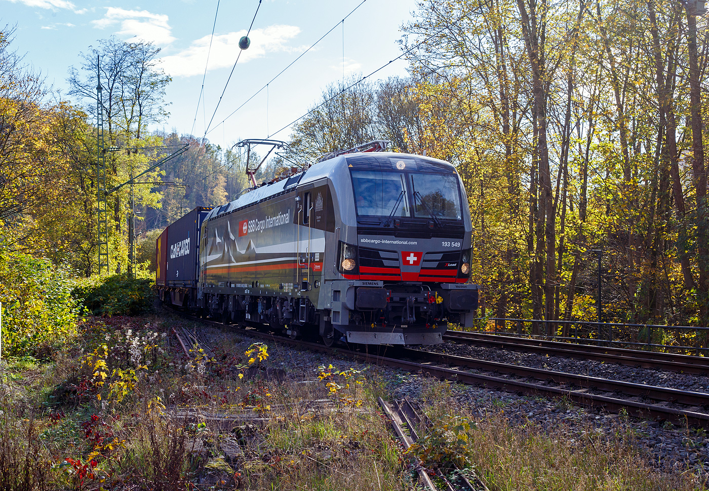 Ein fast fabrikneuer SIEMENS XLoad-Vectron, gerade mal 4 Wochen alt.
Die an die SBB Cargo International AG vermietete 193 549 mit XLoad  Duomo Milano  (91 80 6193 549-3 D-SIEAG) der S�dLeasing GmbH, Stuttgart (eingestellt in Deutschland durch Siemens) f�hrt am 29.10.2024 mit einem KLV-Zug, durch Kirchen (Sieg) in Richtung Siegen.

Die Multisystemlokomotive Siemens Vectron MS wurde 2024 von Siemens Mobilitiy in M�nchen-Allach unter der Fabriknummer 23595 gebaut und am 25.09.2024 ausgeliefert. Sie wurde in der Variante A40-1a ausgef�hrt und hat so die Zulassung f�r Deutschland, �sterreich, die Schweiz, Italien, die Niederlande und Belgien (D / A / CH / I / NL / B). Sie verf�gt �ber eine Leistung von 6,4 MW (160 km/h) und ist neben den nationalen Zugsicherungssystemen mit dem Europ�ischen Zugsicherungssystem (ETCS  BL3) ausgestattet. Zudem ist sie mit der neuen Ausr�stungspaket XLoad ausgestattet. 

Das neue XLoad Ausr�stungspaket f�r Vectron:
XLoad ist ein Ausr�stungspaket f�r Vectron, welches k�nftig mitbestellt, aber auch bei bereits ausgelieferten Vectron Loks nachger�stet werden kann. Das Feature verbessert die Reibwertausnutzung und erm�glicht dadurch h�here Anh�ngelasten. Zudem reduzieren die Fahreigenschaften, die das Feature bewirken, den Verschlei� von Rad und Schiene.

Aktuell sind die Schweizer Vectron-Lokomotiven (SBB Cargo und BLS Cargo) in der Regel in Doppeltraktion unterwegs. Die Steigungen und Rampen der Schweizer Berge sind vor allem bei schlechten Witterungsbedingungen nicht ohne. Eine Lokomotive muss auch bei geringerem Schlupf gen�gend Traktion auf die Schienen bringen, um alle G�ter sicher und zuverl�ssig ans Ziel zu bringen. Ein effizienter Weg aus dieser �Misere� ist die f�r Vectron entwickelte Zusatz-Funktion �XLoad�. Den erfolgreichen Beweis trat eine SIEMENS Testlokomotive im Fr�hjahr 2022 bei der SBB Cargo International und bei der BLS Cargo eindr�cklich an. 

F�r SBB Cargo International bewies die Test-Lokomotive am B�zberg und f�r BLS Cargo an der Nordrampe des L�tschbergs ihre enorme Zugkraft. 
Vectron meisterte im Fr�hjahr 2022 die lange 12‰-Steigung des B�zbergs mit einer Anh�ngerlast von 2.000 Tonnen bravour�s. Bei den n�chtlichen Testfahrten zeigte sich eindr�cklich die enorme Zugkraft der Lokomotive. 

Am L�tschberg wurden bei der BLS Cargo steigungsm��ig noch ein paar Promille draufgepackt. Mit 1.020 Tonnen im Gep�ck bew�ltigte die Vectron-Lokomotive mit XLoad-Feature die 27‰-Steigung der Nordrampe ebenfalls meisterlich. Und auch diverse Anfahrtsversuche absolvierte der mit dem XLoad-Feature aufger�stete Vectron problemlos. 

So bestellte die S�dLeasing GmbH (Stuttgart) im Auftrag der SBB Cargo International j�ngst 20 Vectron Lokomotiven mit XLoad bei SIEMENS.
