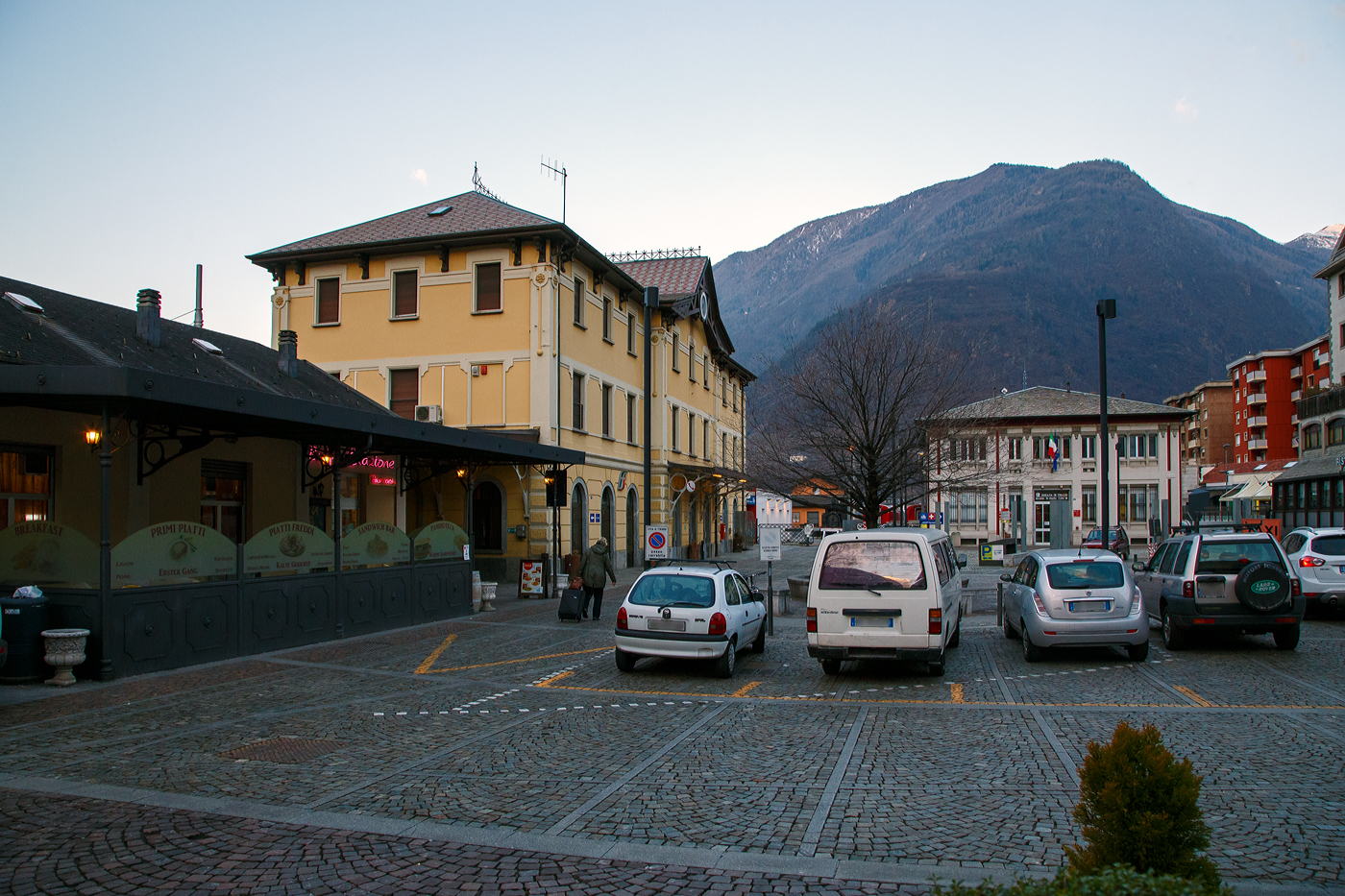 Links der das Empfangsgeb�ude vom FS Bahnhof Tirano an der normalspurigen Bahnstrecke Sondrio–Tirano (RFI 26 / Kursbuchstrecke 180) der Ferrovie dello Stato (FS) hinten rechts der RhB Bahnhof Tirano der meterspurigen Berninabahn der Rh�tischen Bahn (RhB) am fr�hen Morgen vom 20 Februar 2017. 

Beide Bahnh�fe sind jeweils die Endbahnh�fe ihren Strecken. F�r die Berninabahn fungiert er als Grenzbahnhof, zudem handelt es sich um die einzige RhB-Station, die nicht in der Schweiz liegt, sowie um die tiefstgelegene RhB-Station.

Der Bahnh�fe sind auf einer Seeh�he von 429 Metern, der FS Bahnhof wurde 1902 gemeinsam mit der neu erbauten Strecke von Sondrio er�ffnet. Die Anordnung des Empfangsgeb�udes in Seitenlage war eine Bauvorleistung f�r die damals projektierte aber nie realisierte Weiterf�hrung in Richtung Bormio. Im Juli 1908 wurde dann der RhB Bahnhof er�ffnet. 